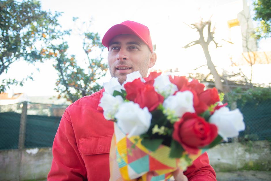 A male florist dressed in a red jacket and matching red cap is holding a vibrant bouquet of fresh flowers outdoors, with a background of trees, a fence, and a bright, sunny sky. The bouquet features a