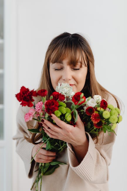 A woman with light brown hair and a gentle expression is holding a vibrant bouquet of fresh flowers close to her face. The bouquet includes red carnations, pink flowers, white hydrangeas, and green bu