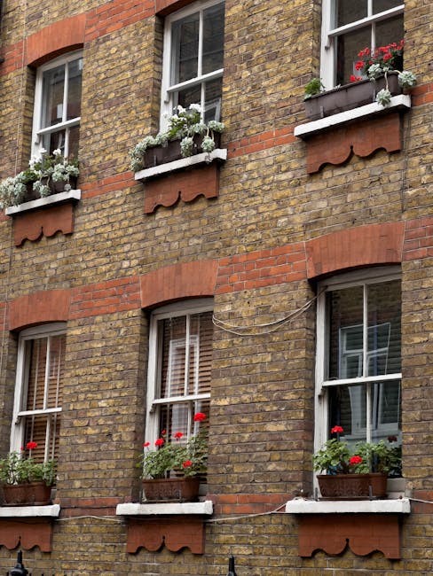 The image depicts a brick building with three rectangular windows, each adorned with white window sills and flower boxes. The flower boxes contain vibrant red geraniums, adding a splash of color again