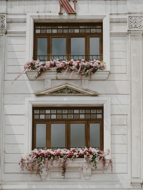 A floral arrangement featuring a lush, cascading bouquet of pink and white flowers, including roses and bougainvillaea, is draped over a white window box on a historic white stone building facade. The