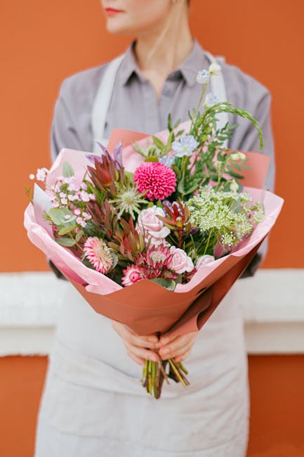 A person dressed in a light grey shirt and aprons holding a large, colorful bouquet of fresh flowers, wrapped in pink and brown paper, against an orange background. The arrangement features pink gerbe