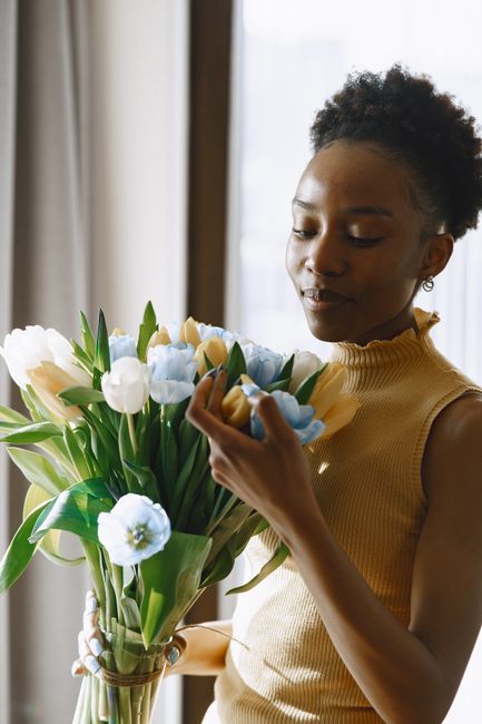 A young woman with dark curly hair, wearing a sleeveless mustard yellow top, is holding a large bouquet of fresh flowers in a clear glass vase. The bouquet includes pale blue, cream, and white tulips 