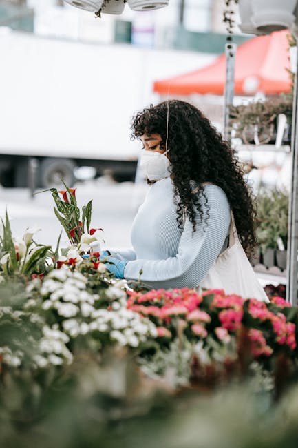 A woman with long, curly dark hair wearing a white face mask, a white sweater, and blue gloves is selecting flowers at an outdoor market stall. She is holding a single red-tipped white lily, with seve