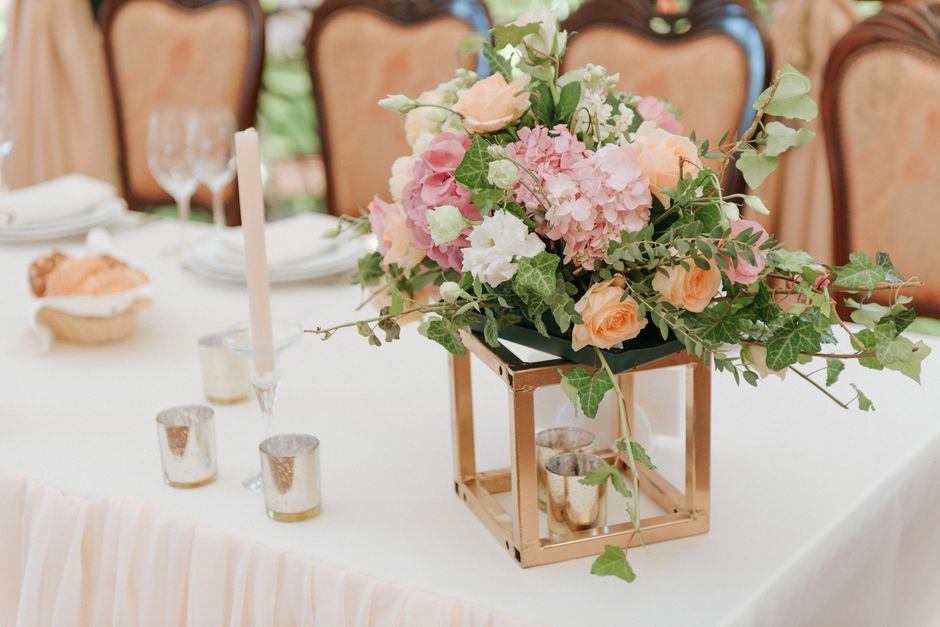 A floral arrangement featuring pink hydrangeas, peach roses, white lisianthus, and green ivy leaves, styled in a black rectangular vase set on a rose gold geometric stand. The bouquet is displayed on 