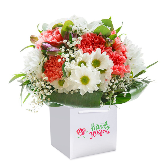 Standing proudly within a pristine white square gift bag, this lushly arranged bouquet brims with the welcoming hues and scents that define New Addington's local flower scene. Soft coral and peach carnations, their frilled petals creating rich textural depth, mingle with vibrant red blooms at the heart of the arrangement. Popping with cheerful contrast, crisp white daisies-each with a sunny yellow-green center-are nestled throughout, brightening the display with their familiar optimism. White sprigs of baby's breath float delicately in the spaces between, creating a gentle, airy veil around the striking blooms. Adding intrigue, hints of tightly closed purple buds nod gently from the left side, suggesting a promise of continued unfolding beauty-much like the well-tended gardens along Lodge Lane. Encircling the flowers, abundant deep green foliage forms a graceful, glossy frame, the broad leaves gathering at the base and fanning elegantly upwards. The entire composition forms a generous, rounded dome, balancing colour, form, and freshness. The bouquet is presented in a smart, modern gift bag-its wide ribbon handles and sturdy structure making local delivery across New Addington effortless and stylish. Set against a flawless white backdrop, the vibrant tones and structural artistry of the bouquet stand out, making it an impeccable choice for milestones, heartfelt gestures, or to simply bring a moment of local colour and seasonal joy into any home.