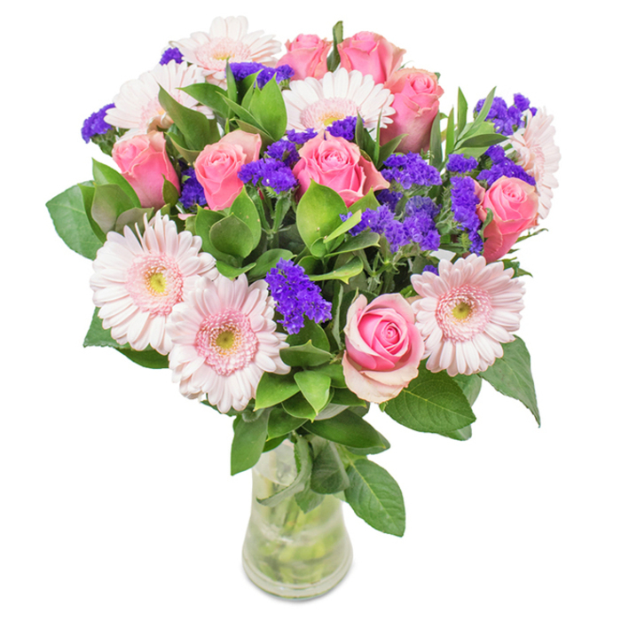 Elegant and vibrant, this floral arrangement from a New Addington florist is a feast for the senses. At its core, a gathering of approximately eight classic pink roses draw the eye, some still curled in tight spirals while others reveal their opulent blooms. These are nestled amidst six pale pink gerbera daisies, their sheer, radiating petals forming soft halos that frame the heart of the bouquet. Clusters of textured, vivid purple statice wind between the larger blossoms, injecting depth and colour contrast, reminiscent of children's laughter echoing through New Addington's parks on a sunny day. Lush, verdant greenery fills out the design, with arching leaves and crisp textures that conjure the freshness of the local countryside after a spring rain. All these elements are rooted in a clear glass vase, where every stem is visible and water catches the light, reinforcing a sense of honesty and natural beauty emblematic of the area. Whether delivered for a special birthday, an anniversary, Mother's Day, or simply to adorn a welcoming home, this arrangement radiates celebration, joy, and a heartfelt connection to its New Addington origins.